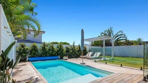 Una piscina rectangular con cubierta de madera, una pérgola y una zona ajardinada delimitada por una valla.