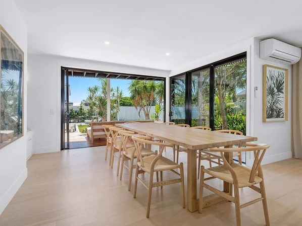 A modern dining area with a long wooden table and chairs, large windows with a view of greenery, and an air conditioning unit on the wall.