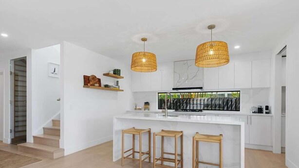 Una cocina moderna con una isla central, taburetes de bar, luces colgantes y una escalera que sube a otro nivel.