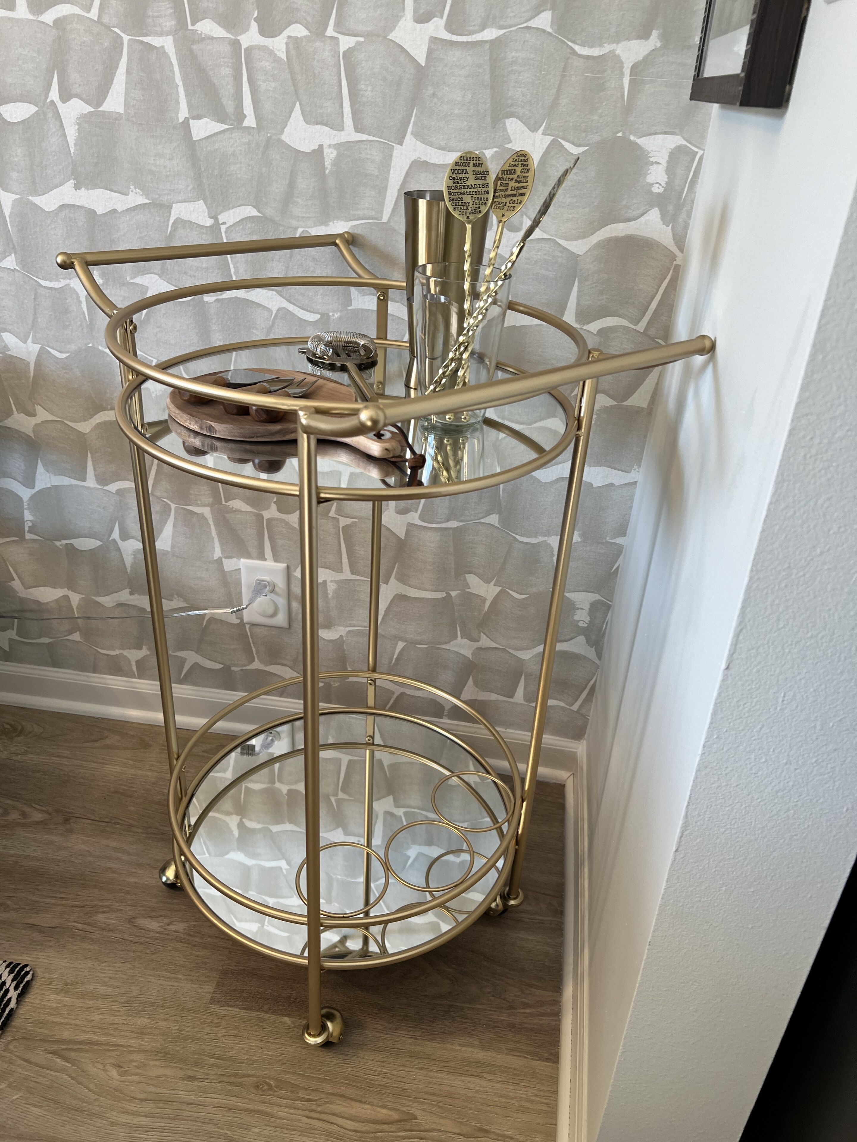 A gold metal stand with a glass top and a small wooden tray, placed against a textured wall.