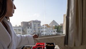 Una persona vestida con una bata blanca sostiene una bandeja con tres recipientes de comida, una botella y una vela, cerca de una ventana con vistas a edificios.