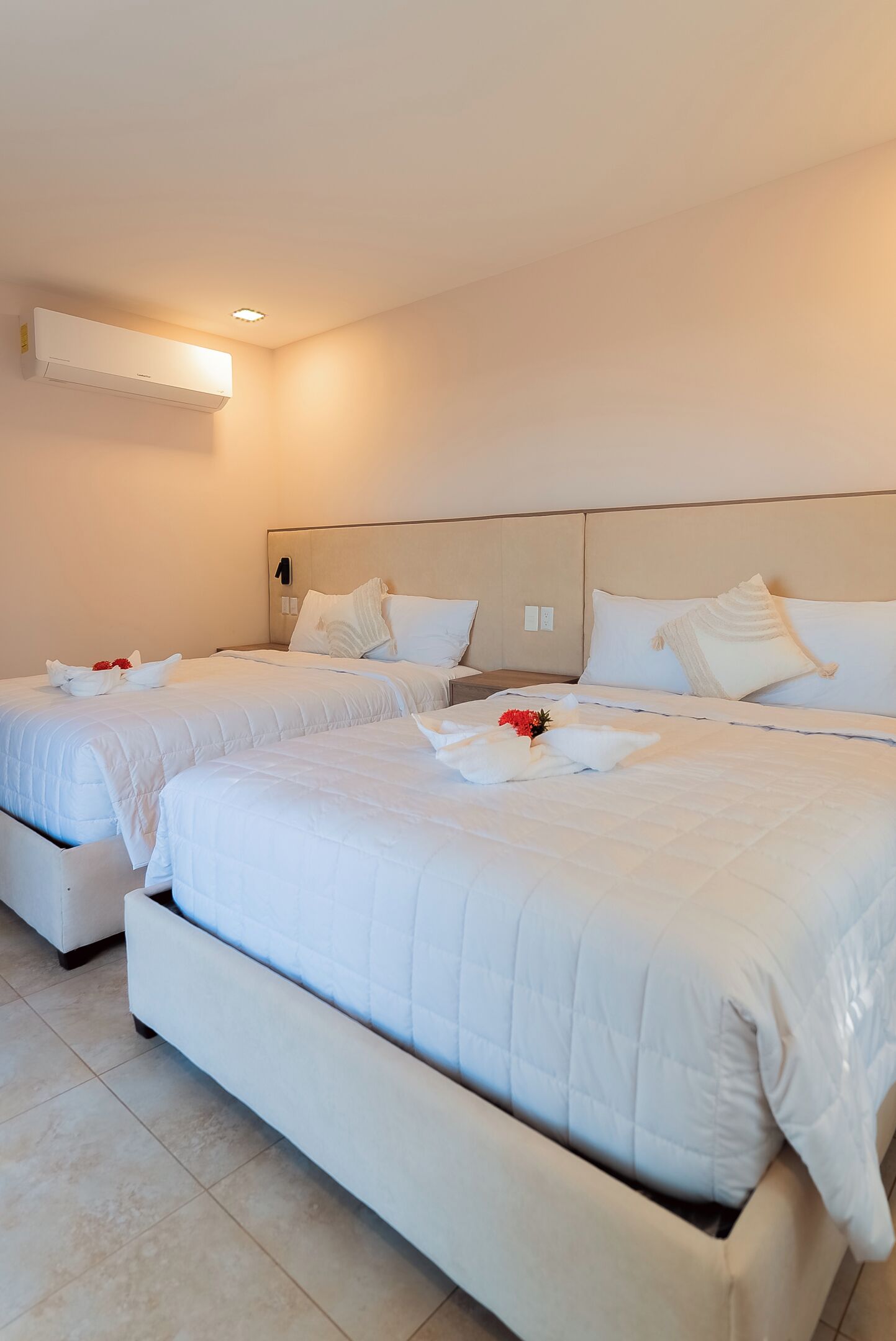 Habitación de hotel con dos camas, cada una con ropa blanca de cama y almohadas, y un aire acondicionado instalado en la pared.