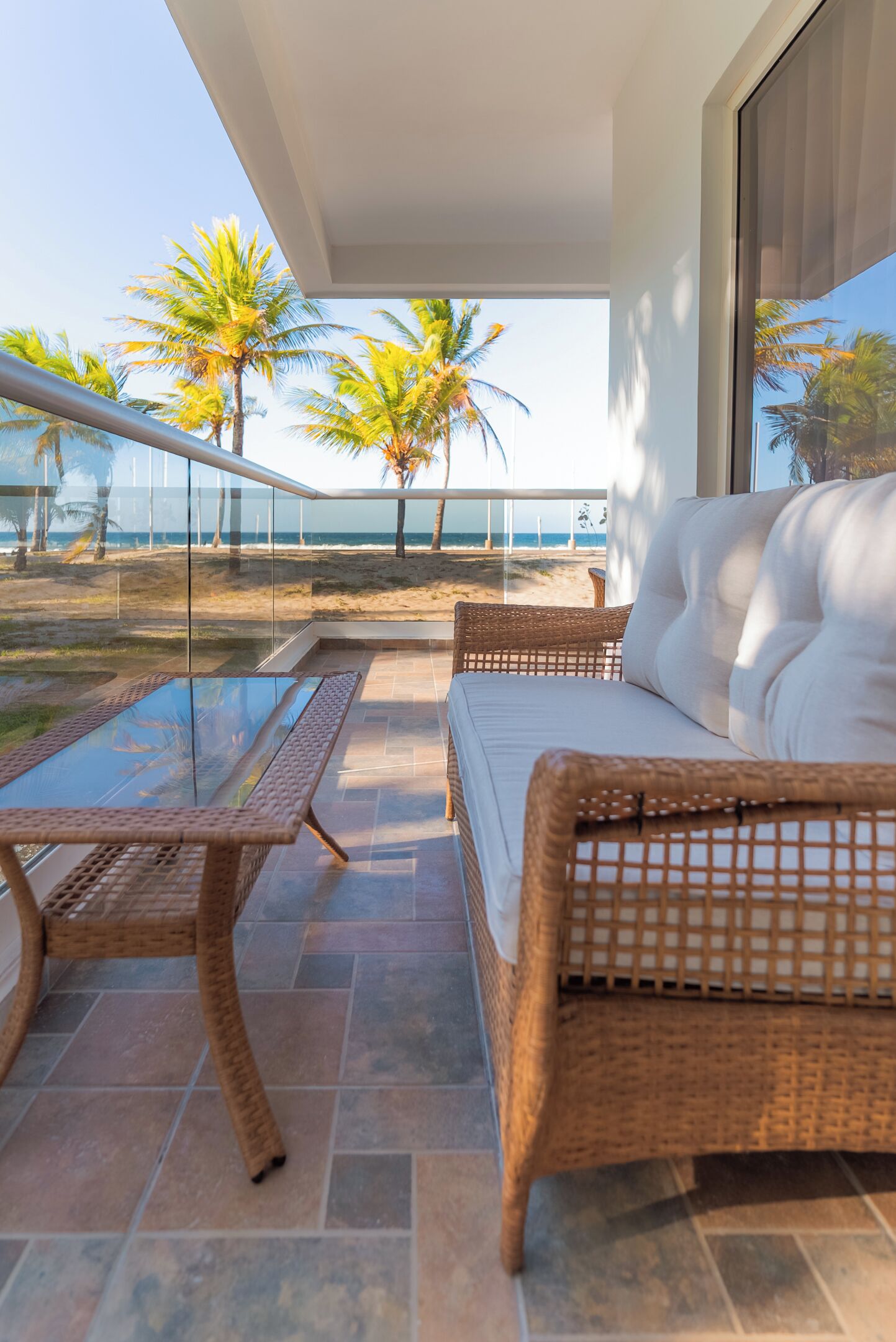 Ein Balkon mit Korbmöbeln, einem Glastisch und Blick auf den Strand und Palmen.