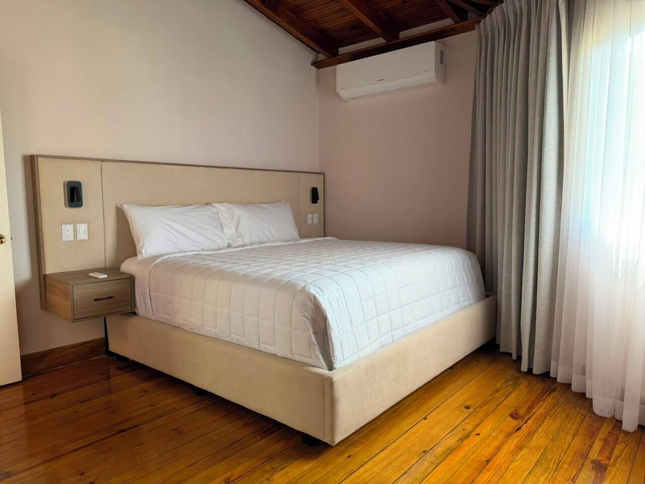 A bedroom with a bed, wooden flooring, a nightstand, and a window with curtains.