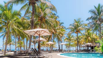 Zona junto a la piscina con sillones de descanso y sombrillas, palmeras y una vista despejada del océano.