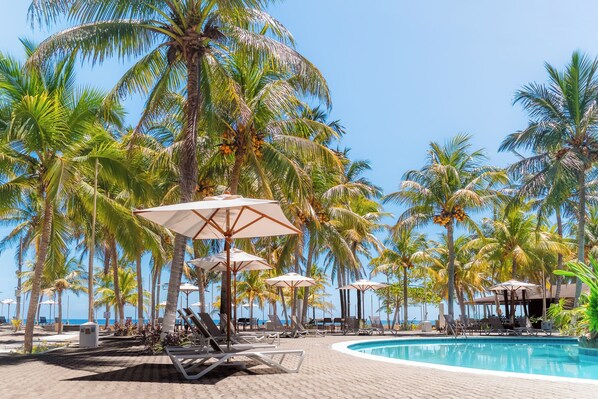 A poolside area with lounge chairs and umbrellas, palm trees, and a clear view of the ocean. - Honduras Shores Plantations (Tela)