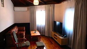Una sala de estar con un sofá, una mesa de centro con un florero, un televisor sobre un mueble bajo y cortinas en las ventanas.