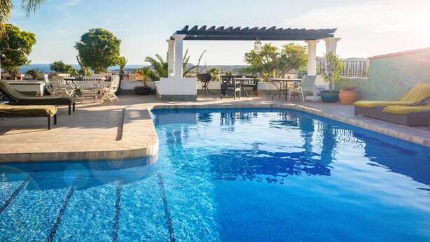 A clear blue pool with lounge chairs and a pergola, surrounded by greenery and a view of the sea.