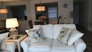 A living room with a white sofa, floral cushions, a table with a lamp, and a kitchen in the background. - Beach lovers paradise!  (Longboat Key)