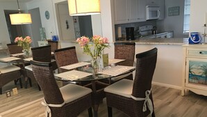 A dining area with wicker chairs and a glass table, a kitchen with white cabinets, and a decorative cabinet with a beach scene. - Beach lovers paradise!  (Longboat Key)