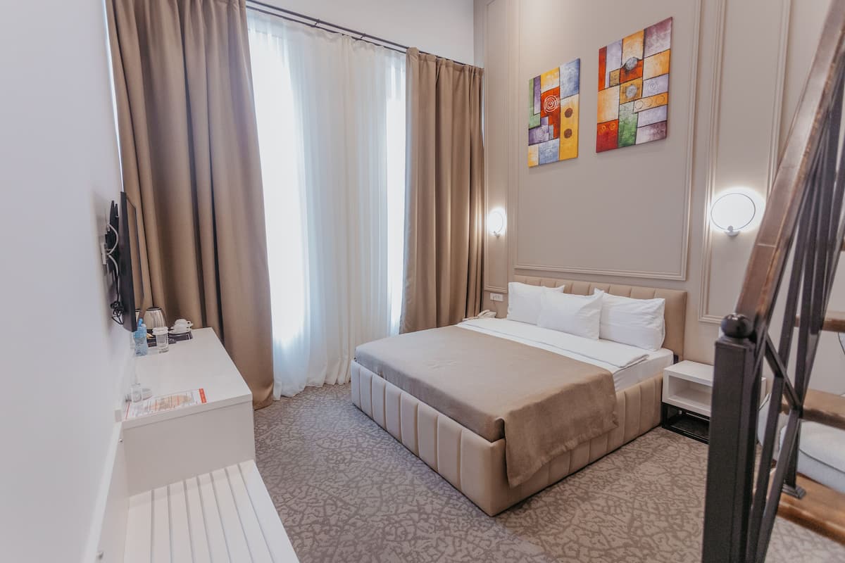 A hotel room with a bed, bedside table, and a desk with a chair. There are curtains, a painting on the wall, and a light fixture above the bed.