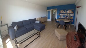 A modern living room with a sectional sofa, a glass coffee table, a dining area with a table and chairs, a bookshelf, and a TV stand. - Appartement Très Agréable Avec vue Dégagée Proche de la Plage et des Commerces (La Baule-Escoublac)