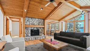 A spacious living room with a stone fireplace, wooden ceiling beams, and large windows offering a view of the outdoors. - Buckatabon Lakeside Retreat. Direct access to snowmobile trails! (Conover)