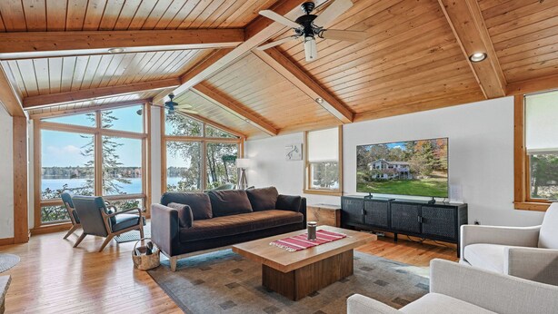 A spacious living room with wooden flooring, a large window overlooking a lake, a ceiling fan, and a television mounted on the wall. - Buckatabon Lakeside Retreat. Direct access to snowmobile trails! (Conover)