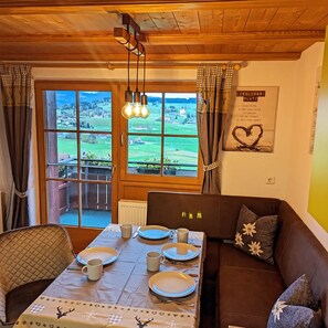 Un coin repas accueillant, avec une table dressée pour quatre personnes, un canapé marron et une vue sur un paysage verdoyant par de grandes fenêtres.