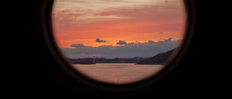 Une vue sur le coucher de soleil à travers une fenêtre circulaire, avec en arrière-plan un plan d’eau, des montagnes lointaines et un pont.