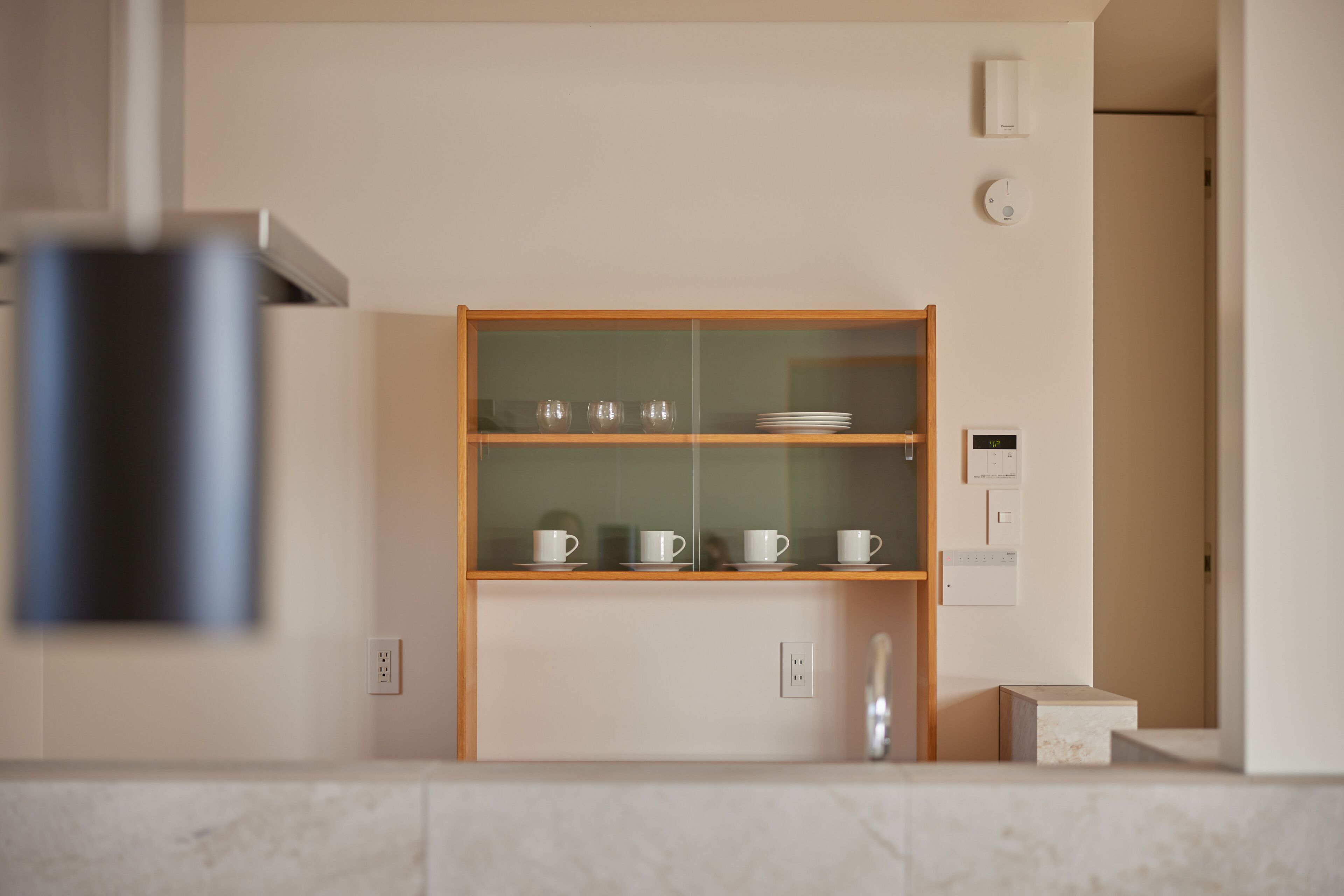 A kitchen with a wooden cabinet containing cups and plates, a white countertop, and a wall-mounted light switch.