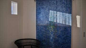 A room with a large blue textured wall hanging, a small window, and a black wicker chair.