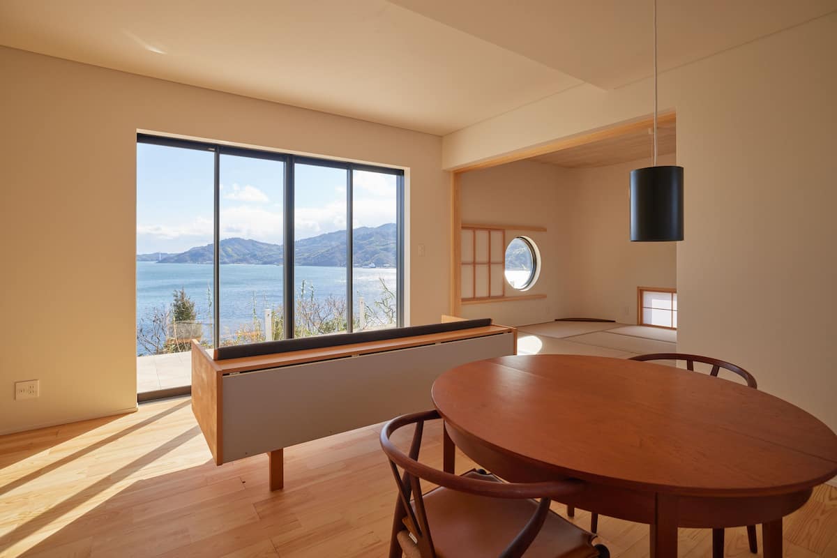 A modern dining area with a wooden table and chairs, large windows offering a view of the sea and mountains, and a minimalist interior design.