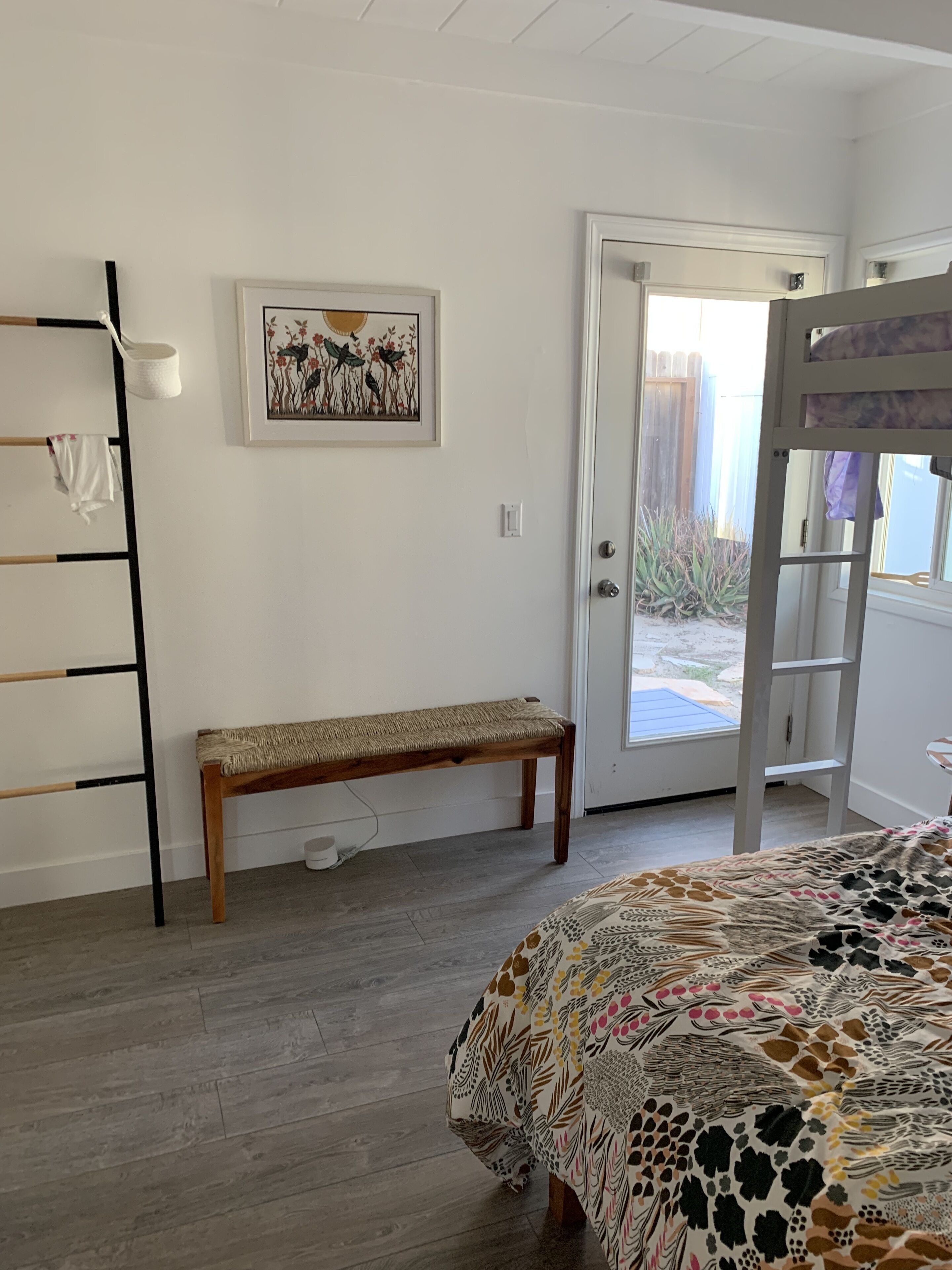 A bedroom with a wooden floor, a bench, a bed with a patterned bedspread, a ladder, a door leading outside, and a framed picture on the wall.