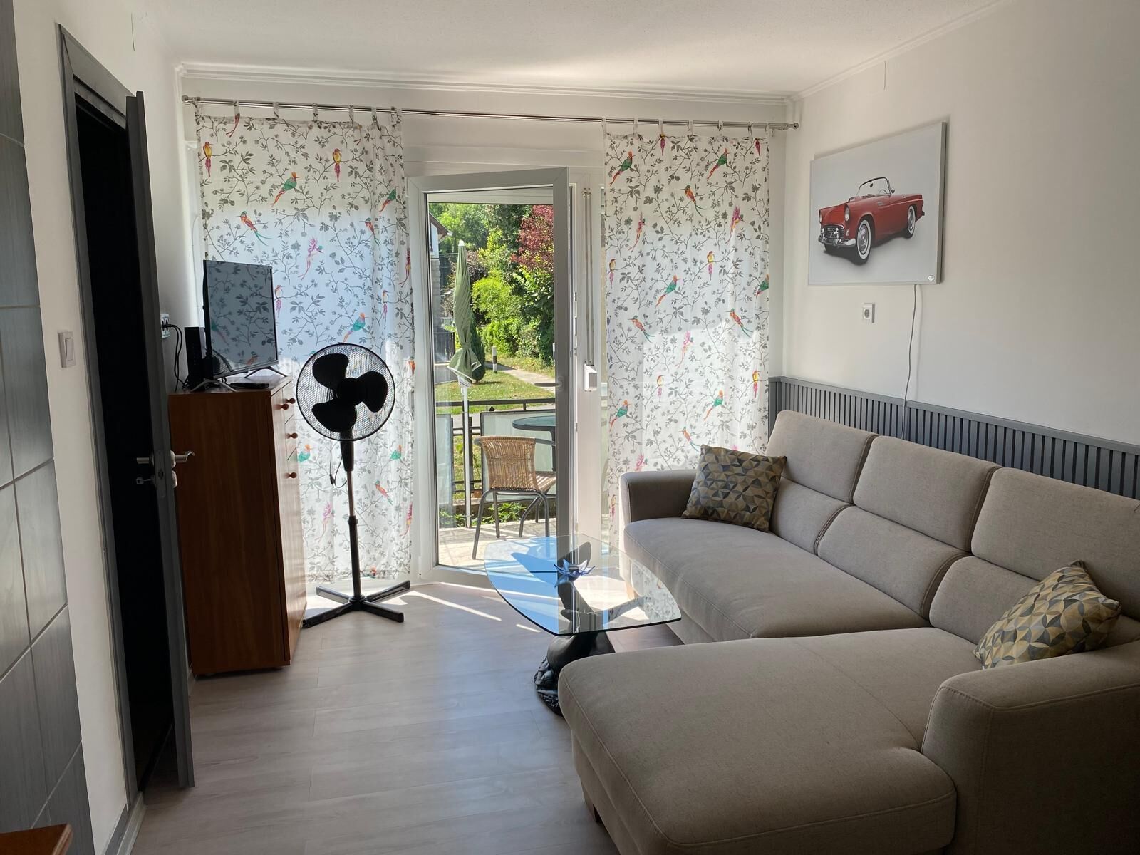 A living room with a sofa, a fan, a table, and a door leading to a balcony.