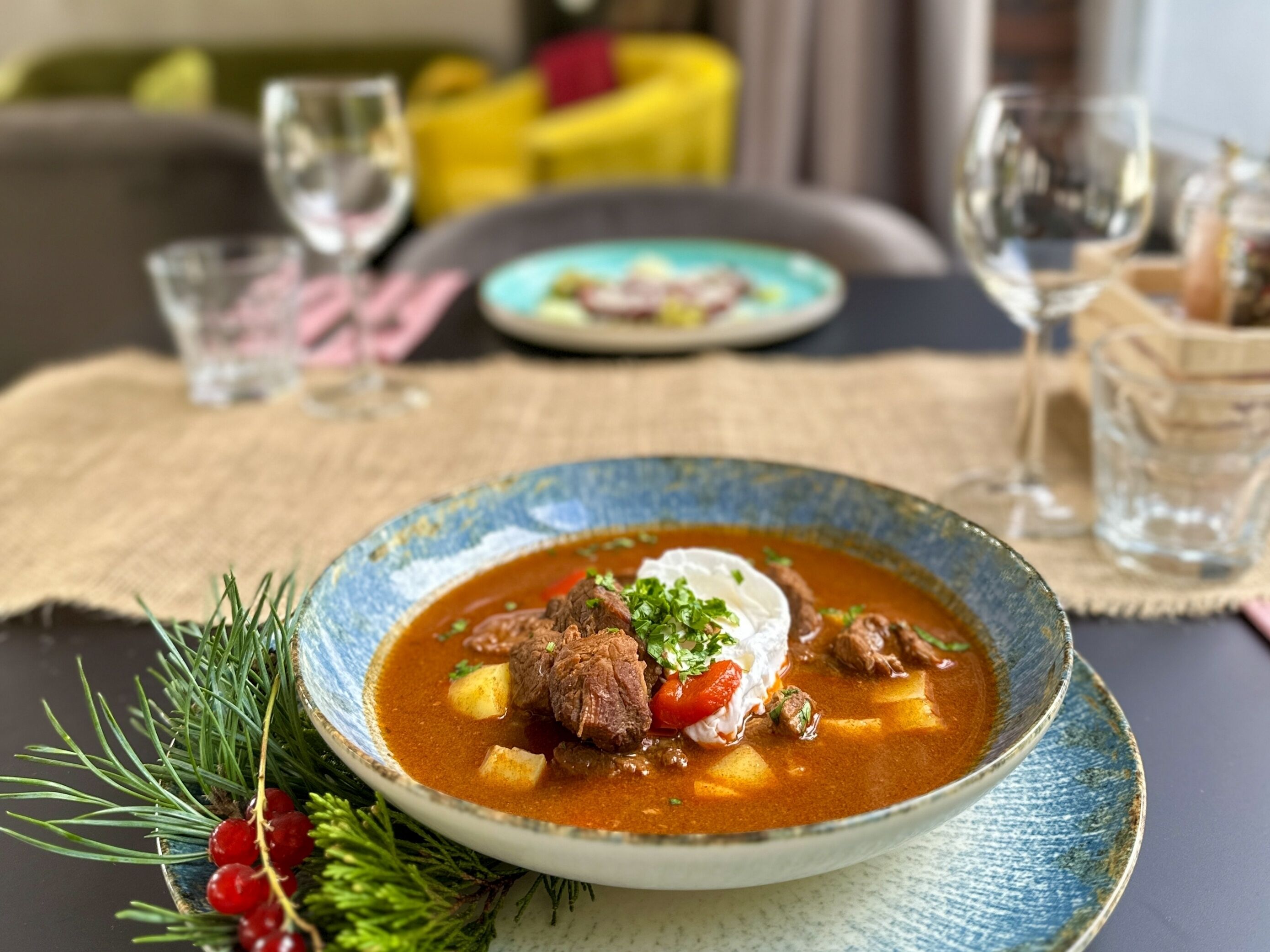 Uma tigela de ensopado com carne, batatas e uma colherada de creme azedo, decorado com ervas, sobre uma mesa com tecido de saco.