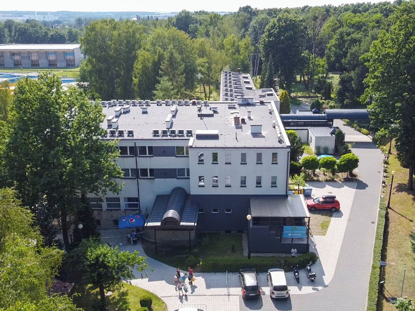 Um edifício com vários andares e telhado plano, cercado por árvores e um estacionamento.