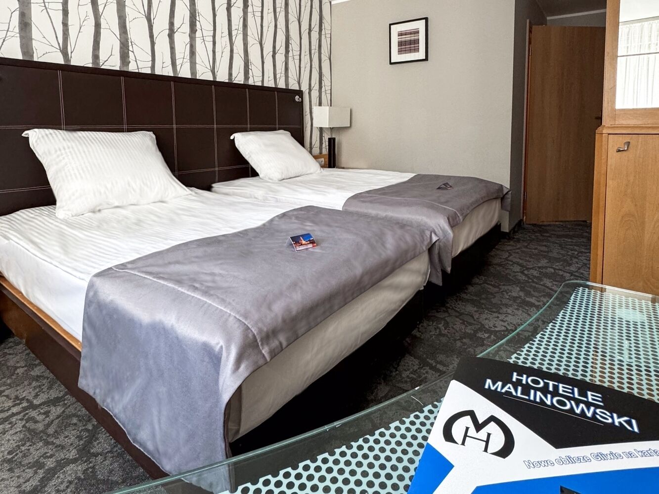 A hotel room with two beds, a wooden headboard, a wall-mounted lamp, and a sign for Hotel Malinowski.