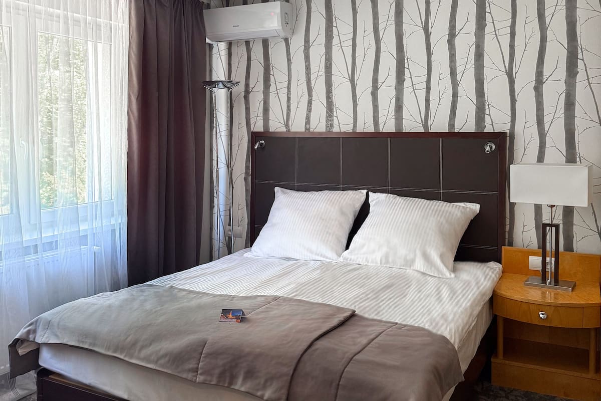 A hotel room with a bed, a nightstand, an air conditioner, and a window with curtains.