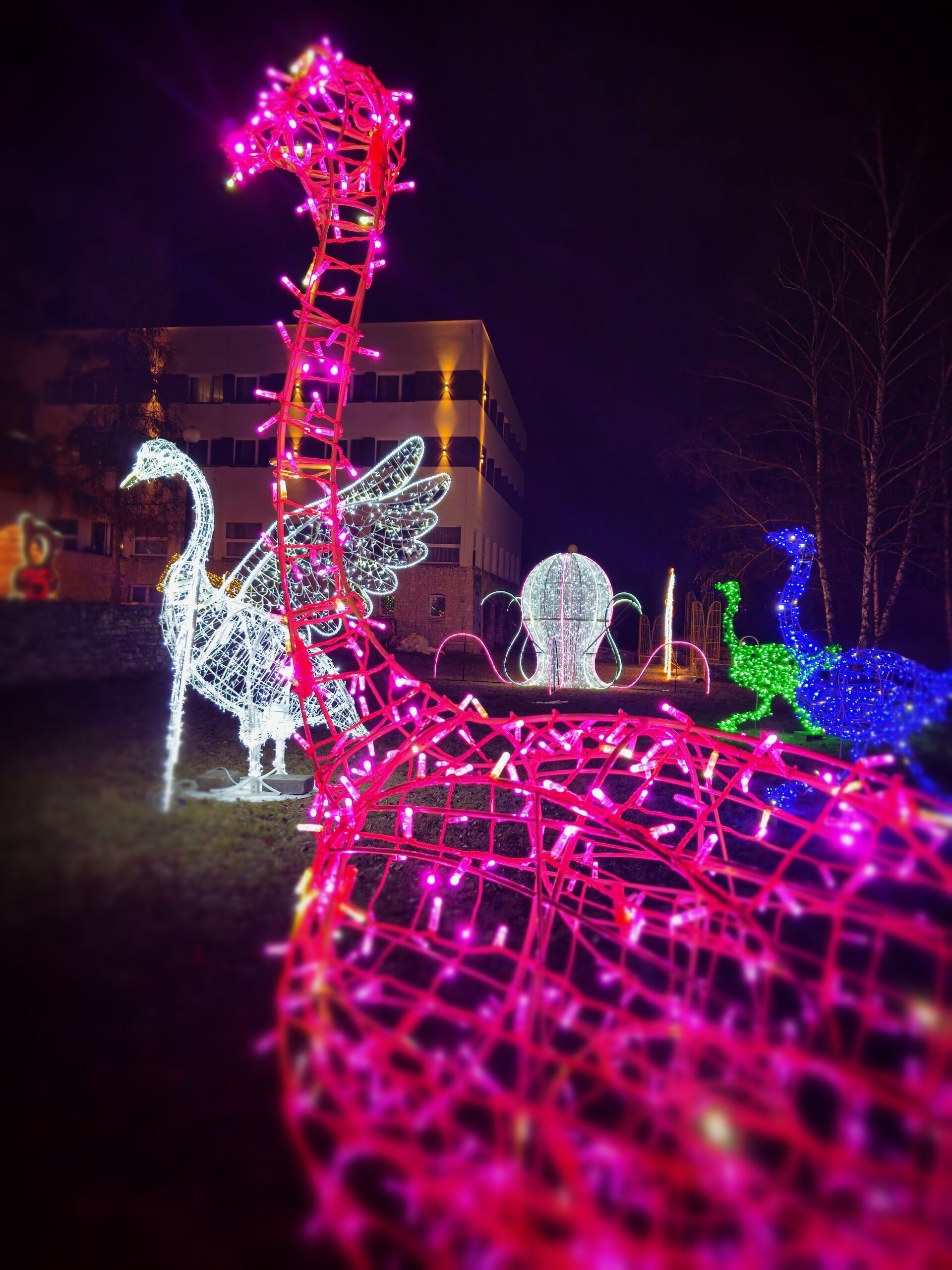 Escena nocturna con estructuras iluminadas, incluyendo un cisne, una mano y una serpiente, con un edificio al fondo.