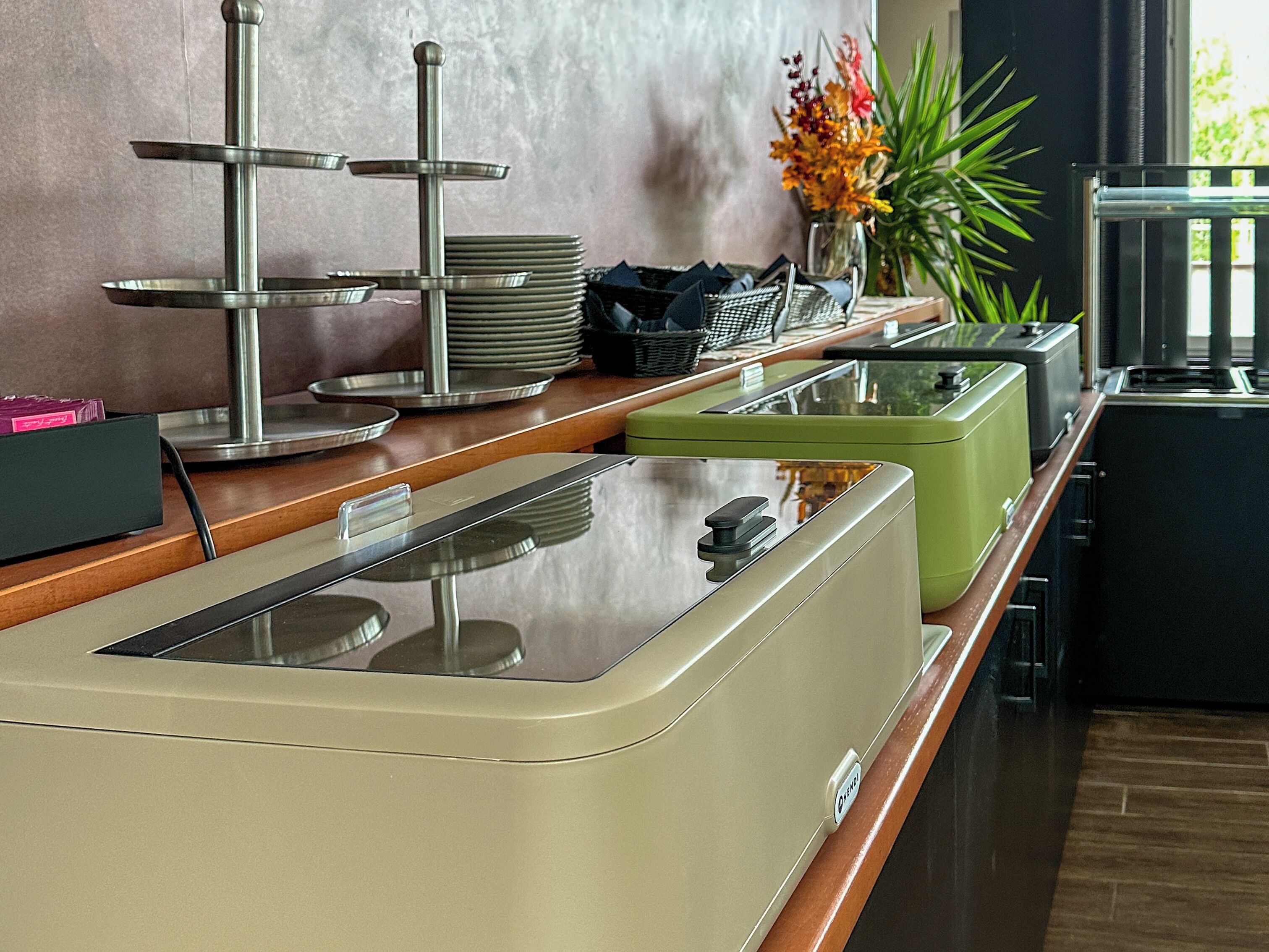 Un espace buffet avec un coin où les clients peuvent se servir eux-mêmes, une pile d’assiettes et un vase rempli de fleurs.