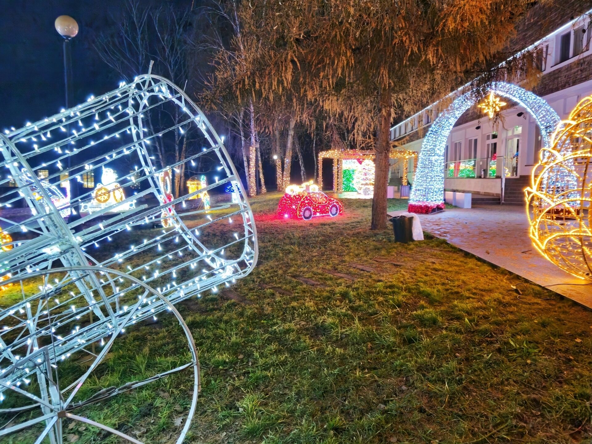Un espacio exterior bien iluminado con luces decorativas, incluyendo una estructura grande iluminada que parece un coche y una exhibición esférica de luces.