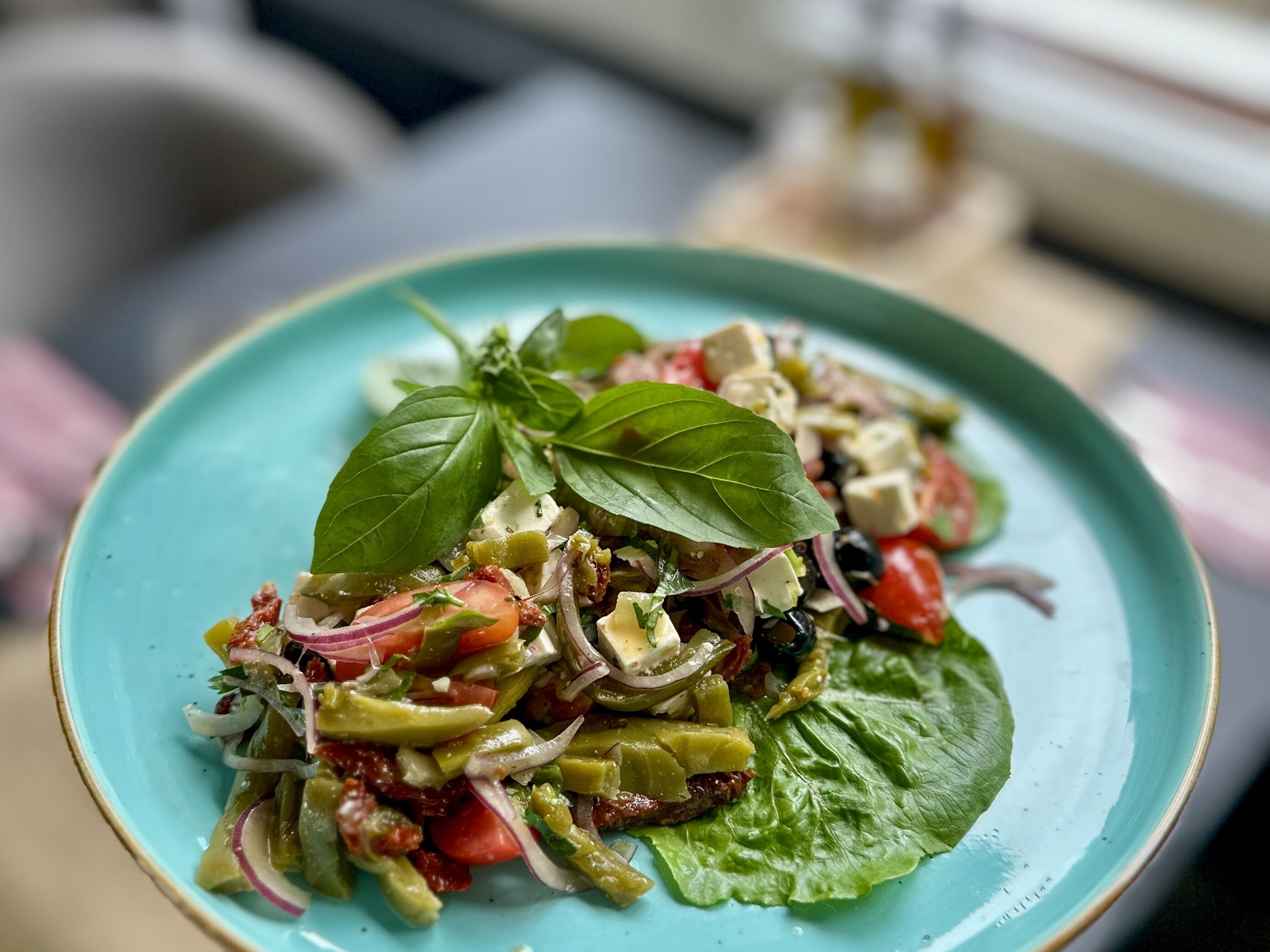 A plate of mixed salad with various vegetables and cheese, garnished with basil leaves, on a turquoise plate.