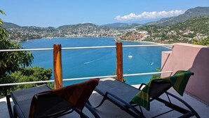Varanda com duas cadeiras, vista para uma cidade costeira com barcos na água e colinas ao fundo.