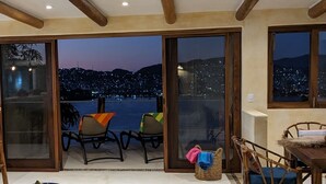 Quarto com uma grande porta de vidro deslizante que dá para uma varanda com cadeiras e vista para a cidade à noite.