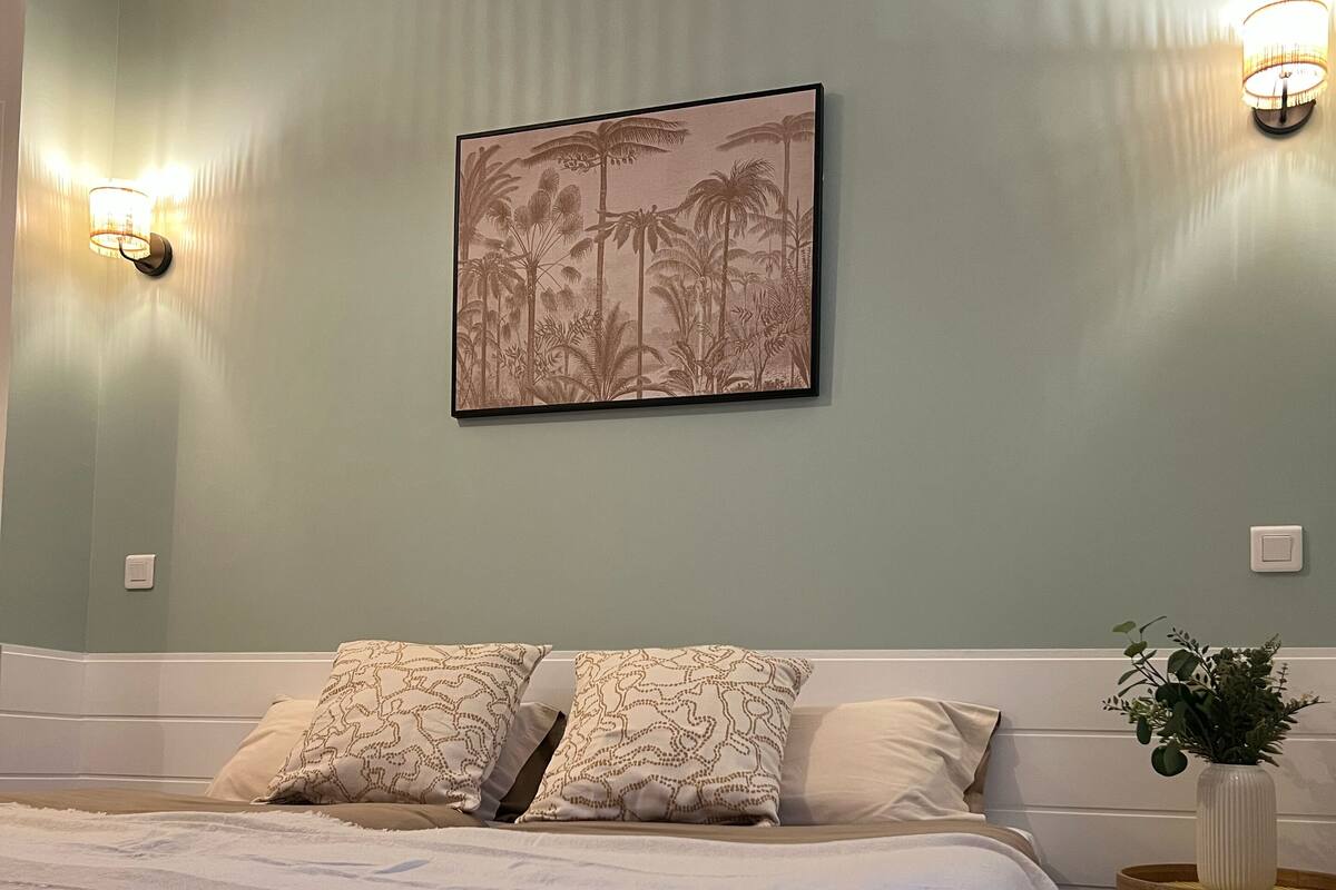 Une chambre à coucher avec un lit, deux lampes fixées au mur, un tableau encadré et une petite table sur laquelle se trouve une plante.