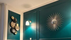 Une chambre avec un mur de couleur turquoise, un miroir rond orné d’un motif en forme de soleil, et un lit avec des coussins à motifs.