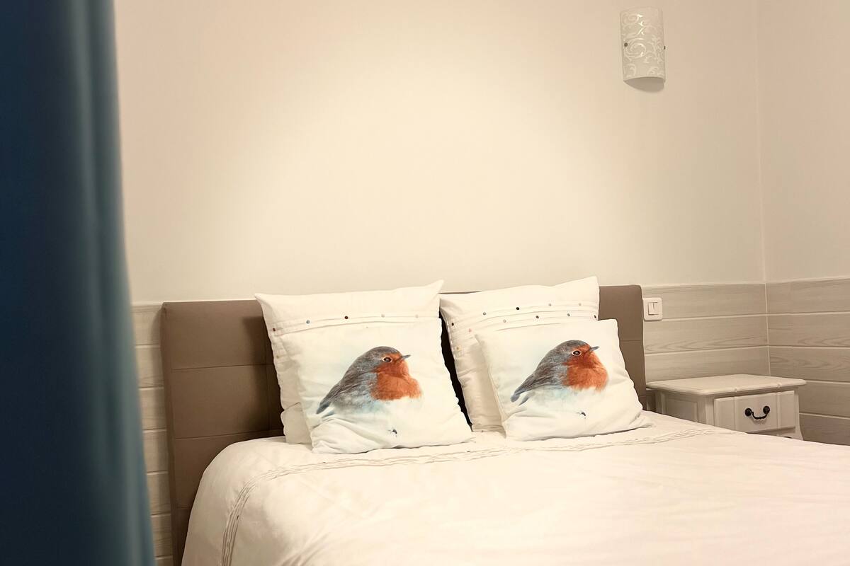 Une chambre à coucher avec un lit, deux oreillers ornés de motifs d’oiseaux, une table de chevet et une commode.