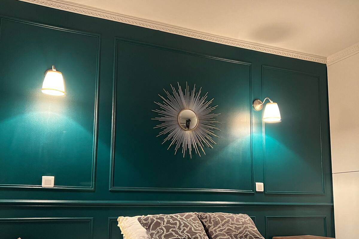 Une chambre avec un mur de couleur turquoise, un miroir rond orné de motifs rayonnants, deux lampes de chevet, un lit avec des oreillers blancs et à motifs, et un plaid vert foncé.