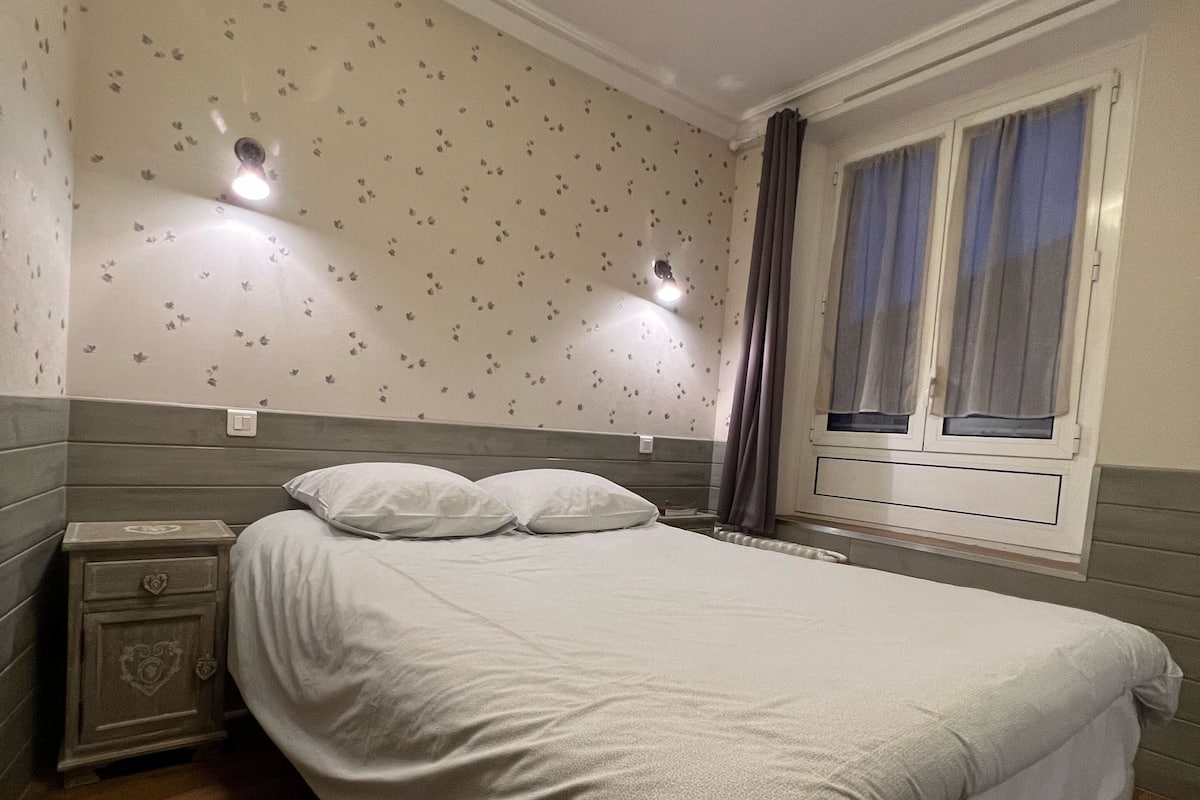 Une chambre à coucher avec un grand lit, deux tables de chevet, une fenêtre avec des rideaux et un mur orné d’un motif floral.