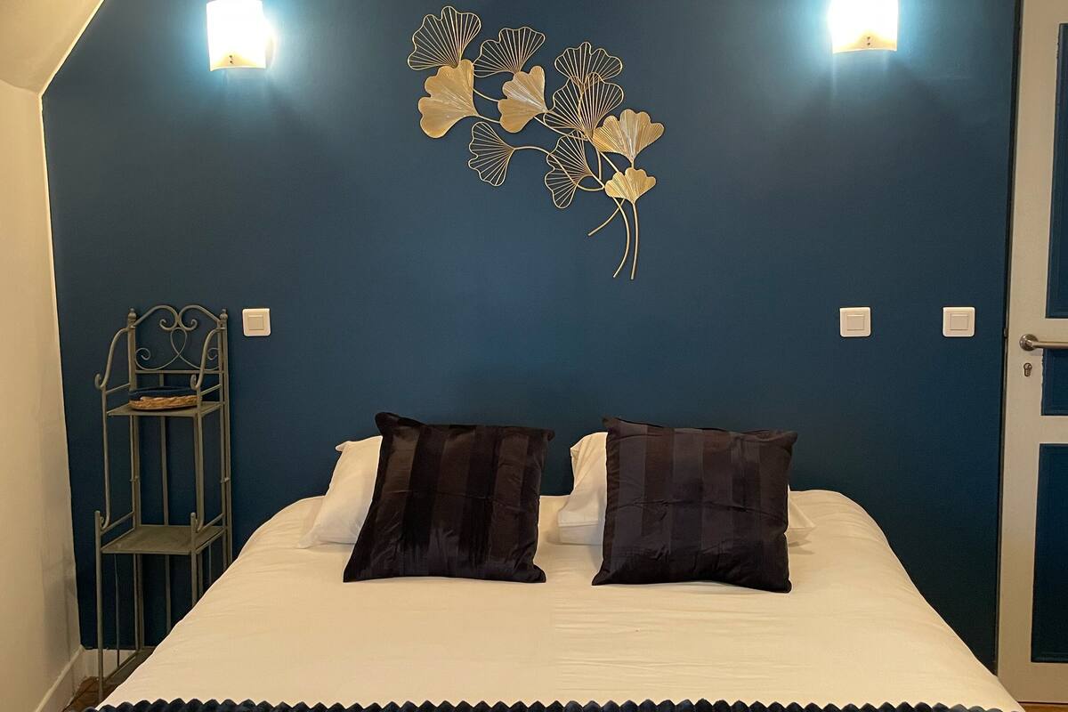 Une chambre à coucher avec un lit, des murs bleu foncé, deux lampes fixées au mur, un élément décoratif mural et une table de chevet avec un panier.