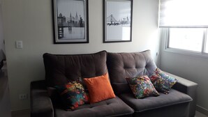 Sala de estar com um sofá marrom escuro, duas almofadas coloridas e duas imagens emolduradas de paisagens urbanas na parede.