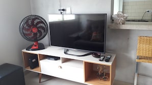 Uma TV de tela plana sobre um móvel branco, com um ventilador vermelho ao lado, um controle remoto e um objeto decorativo na prateleira acima.
