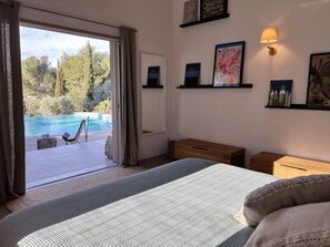 Ein Schlafzimmer mit einem großen Bett, einem Holfnachtkästchen, einem Nachttisch und einer Wandlampe. Ein Fenster mit Vorhängen bietet Blick auf einen Außenpool, und eine Tür führt nach draußen.