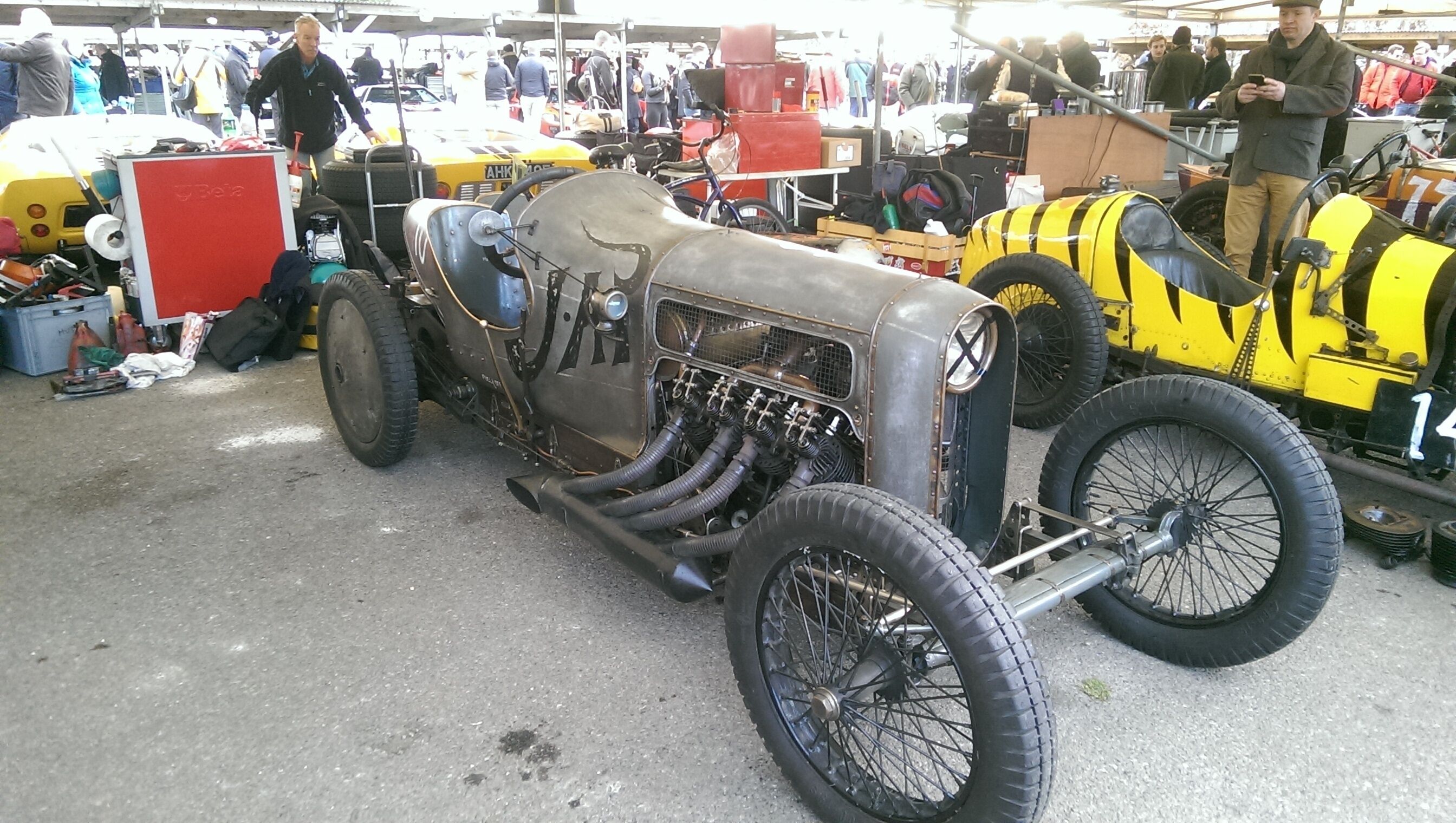 A vintage car with exposed engine parts and a large front grille, parked at an outdoor event with other vehicles and attendees in the background.