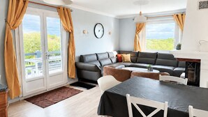 Un salon moderne comprenant un canapé, une table à manger et des chaises, de grandes fenêtres avec des rideaux, une horloge et une cheminée.