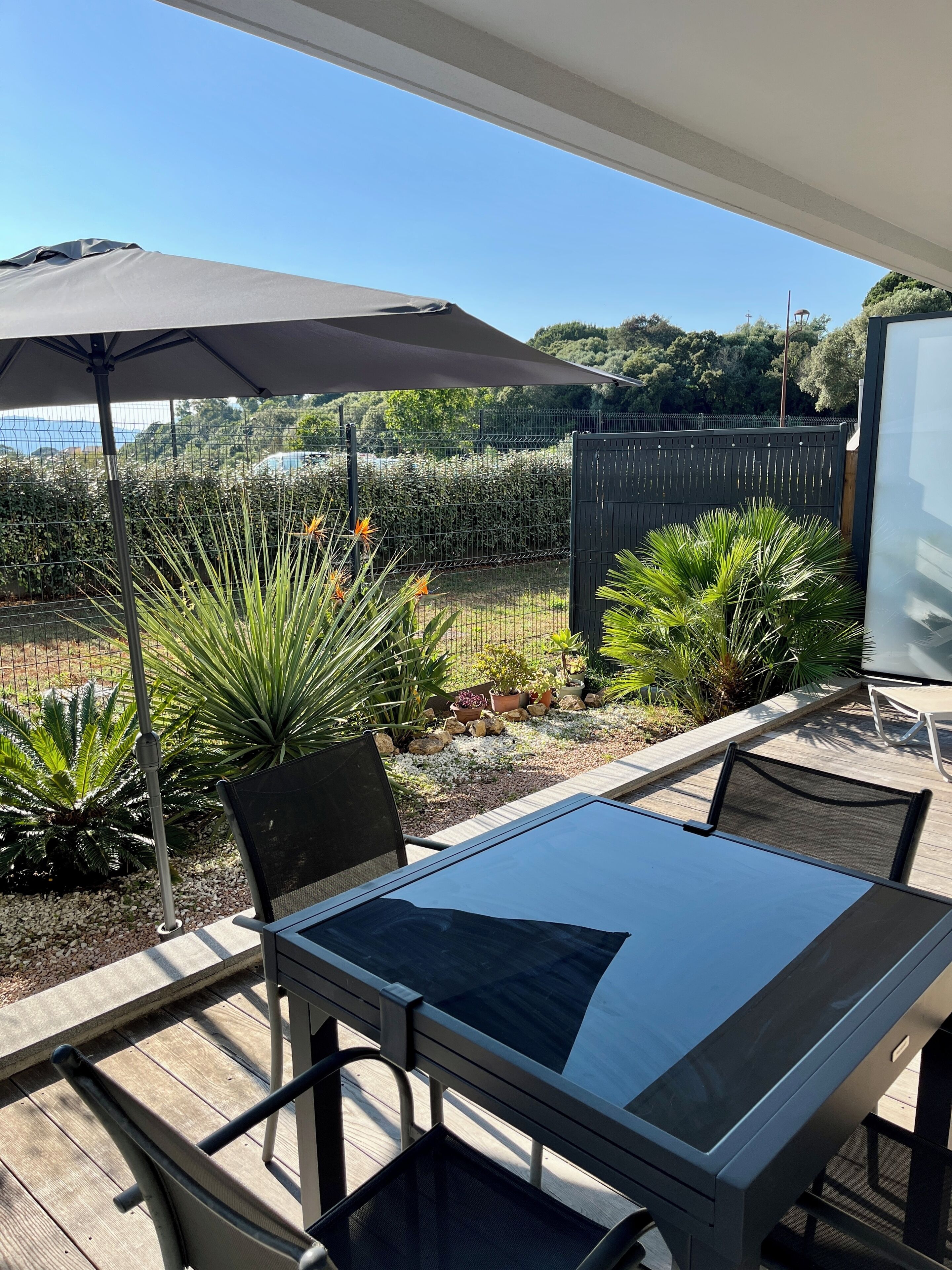 Une terrasse avec une table et des chaises, un parasol, et une vue sur un jardin et une clôture.