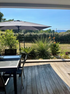 Une terrasse en bois avec une table et des chaises, un grand parasol, et une vue sur un jardin aménagé avec différentes plantes et une clôture.