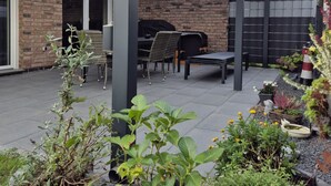 Ein überdachter Patio-Bereich mit Gartenmöbeln, einem Grill und Topfpflanzen.