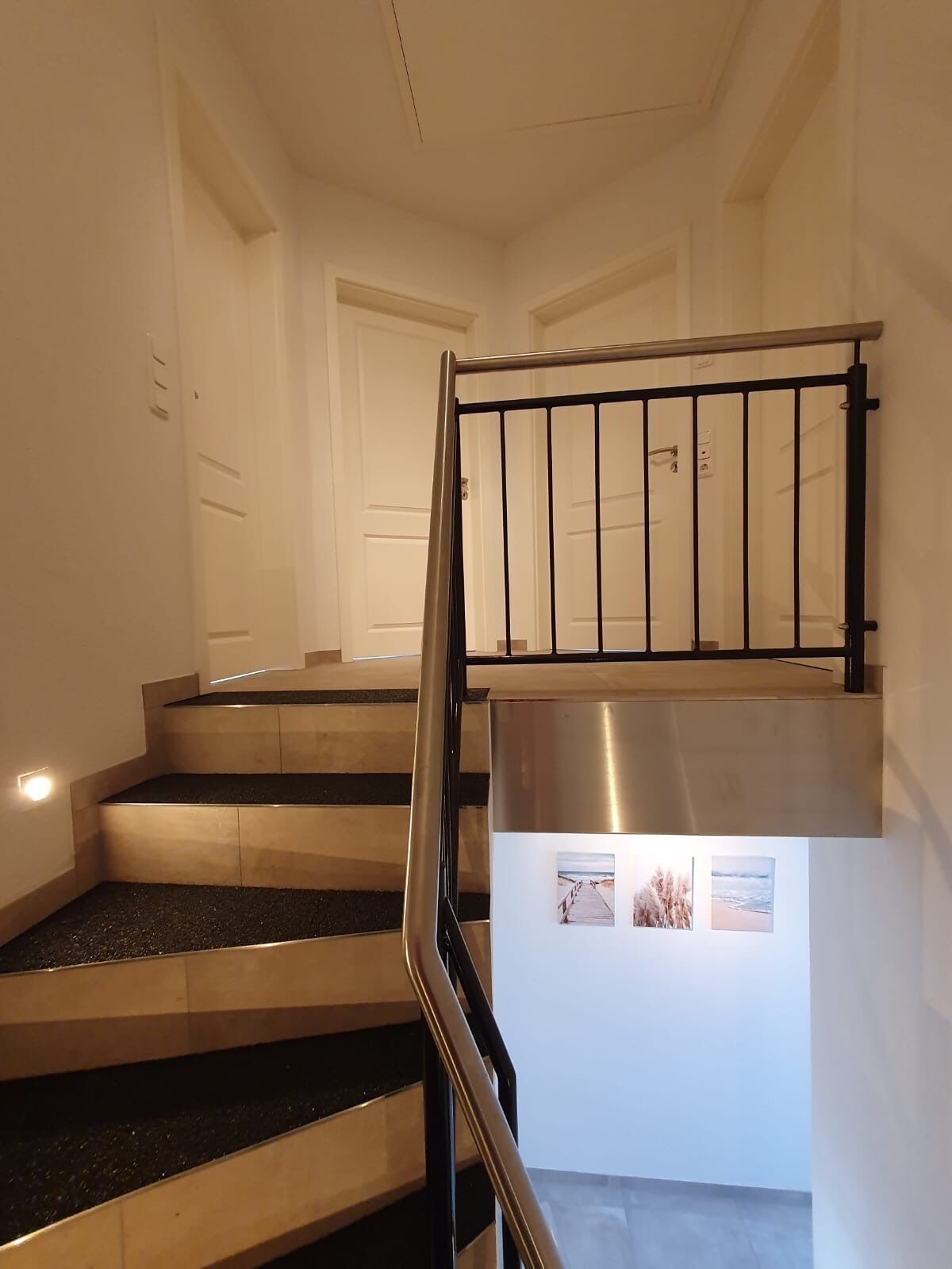A staircase with a metal handrail, a landing with a stainless steel column, and a door in the background.